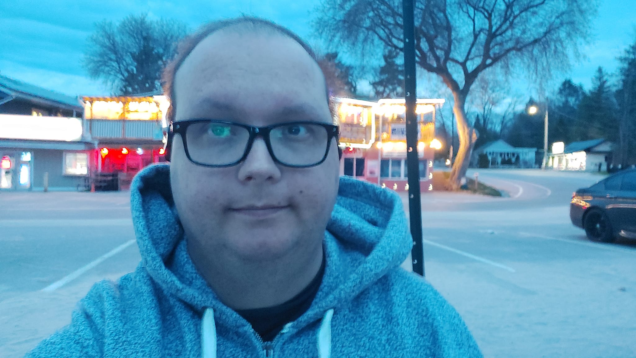 A man wearing a hoodie with very little hair sits at a bench at dusk, a brightly lit bar is behind him